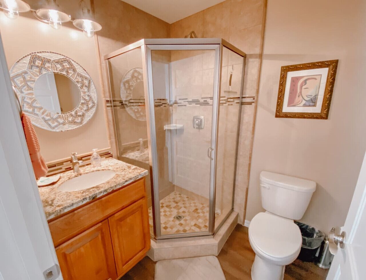 A bathroom with a glass-enclosed shower, a sink with a marble countertop, wooden cabinets, a toilet, and a framed artwork on the wall.