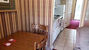 Uma sala de jantar com mesa e cadeiras de madeira, cortina listrada, pequeno espaço de cozinha com eletrodomésticos brancos e tapete vermelho.