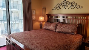 A hotel room with a large bed, a wooden headboard, a nightstand, a lamp, and a window with curtains.