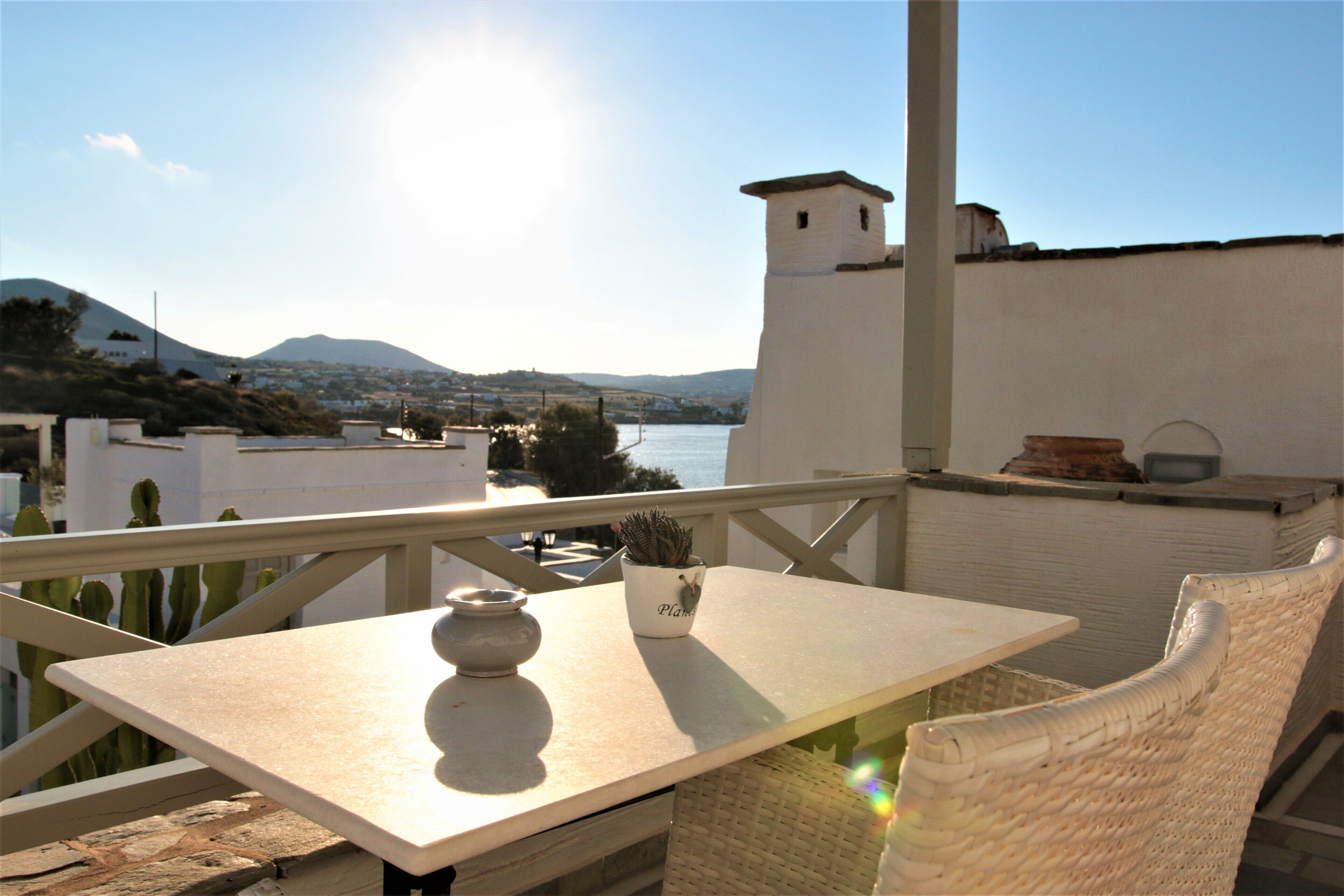 A balcony with a table and wicker chair, overlooking a body of water and distant hills.