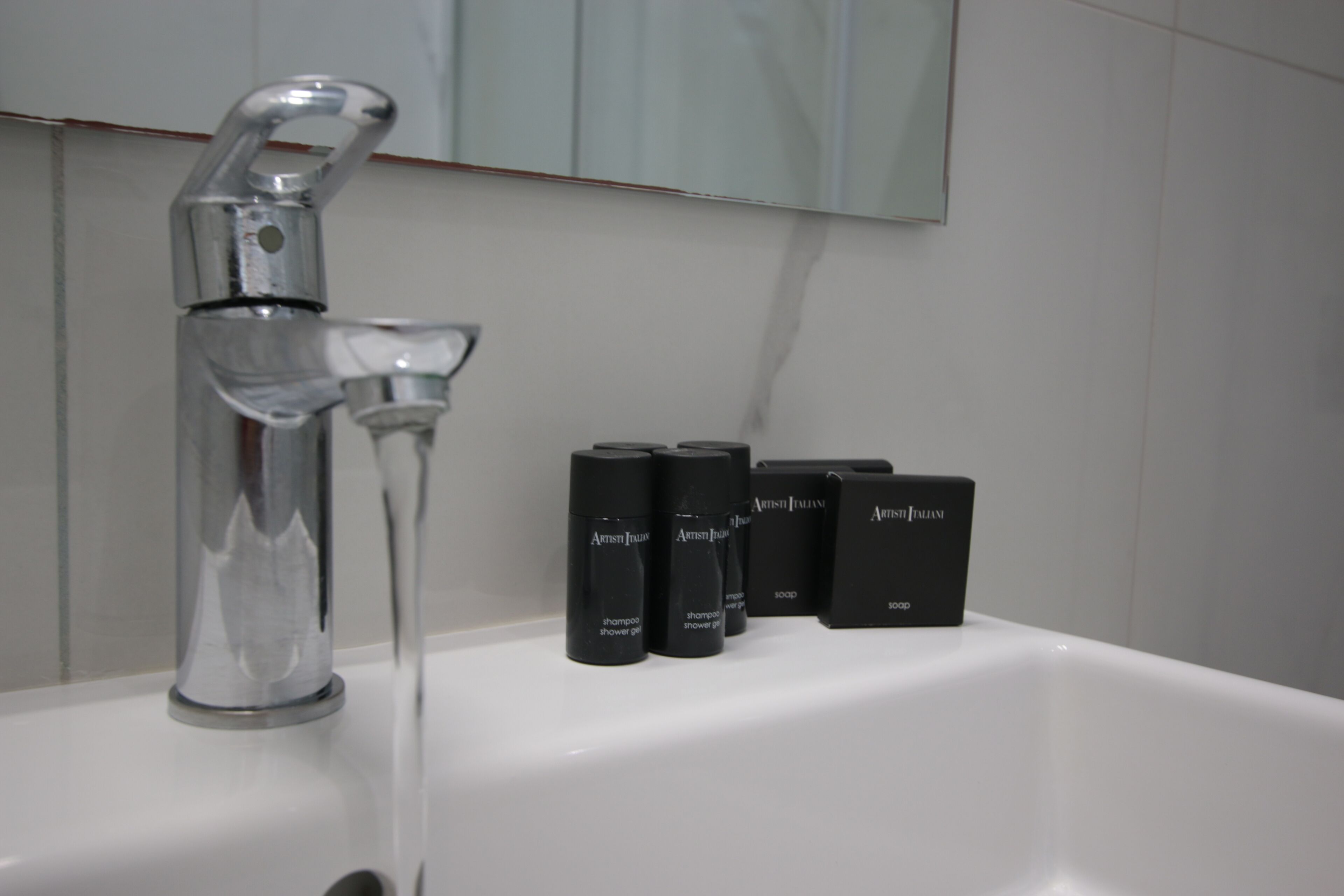 A bathroom sink with a chrome faucet and running water. There are four black bottles labeled 'Artist Italian' on the sink.