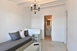 A modern, minimalist room with a grey sofa, a small white table, and a glass cabinet. A chandelier hangs from the ceiling, and a bathroom is visible in the background.