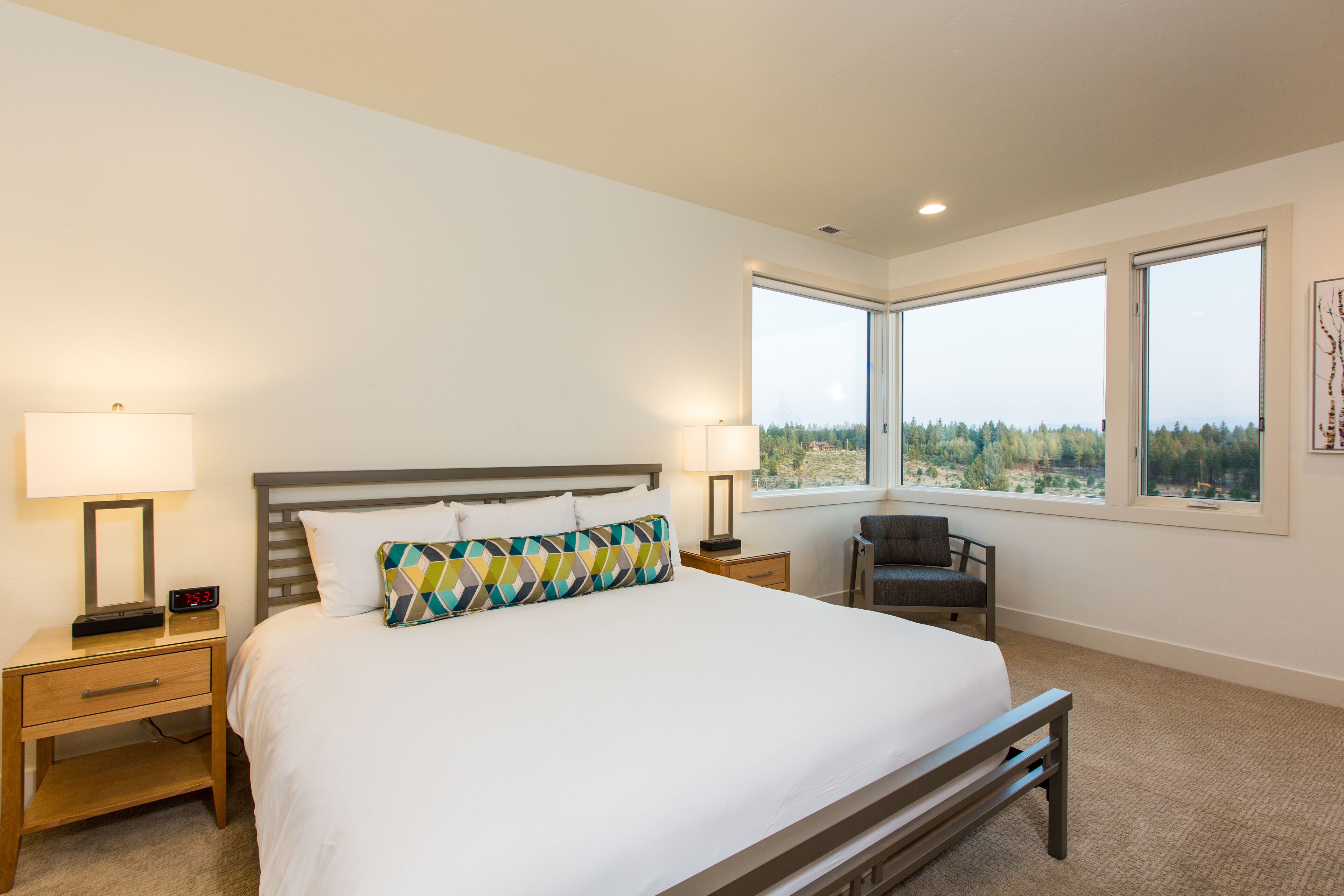 A bedroom with a large bed, two bedside tables with lamps, a chair, and a view of the outdoors through large windows.