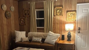 A cozy living room with a brown wooden panel wall, a brown sofa with white cushions, a small table with a lamp, and a patterned rug. - The Charming Country Cottage-Welcomes you and your pet! (La Plata)