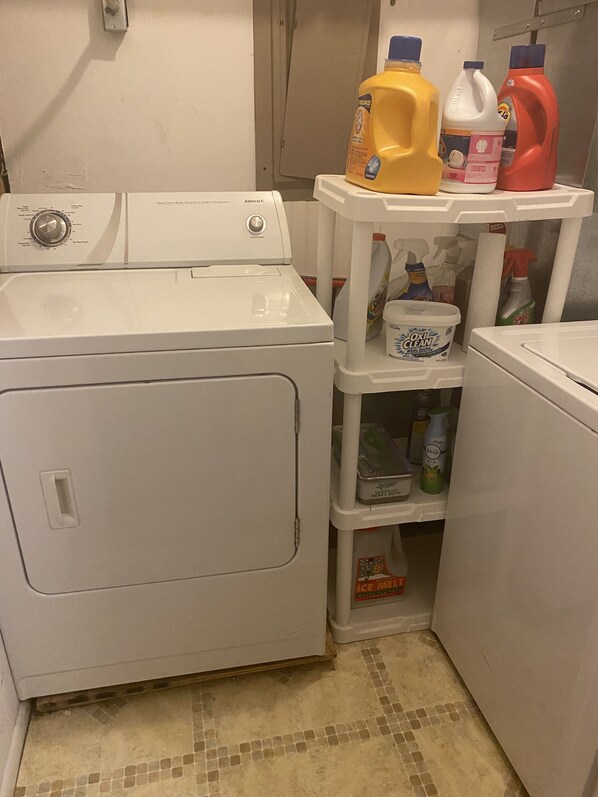 A laundry area with a front-loading washing machine, a shelf with detergent bottles, and a container of oil.