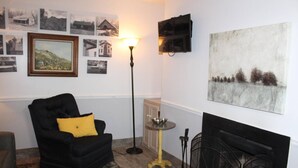 Una sala de estar acogedora con un sillón negro, un cojín amarillo, una chimenea y una colección de fotografías enmarcadas en la pared.