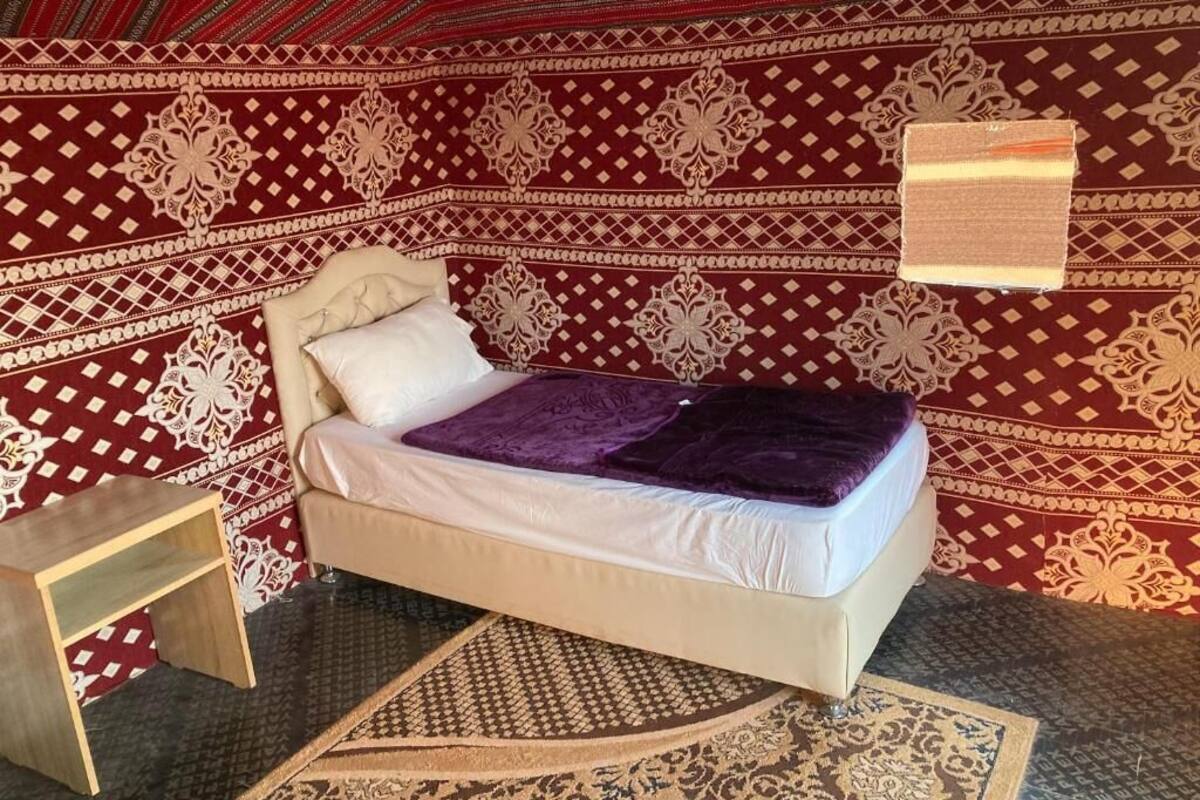 A single bed with a white mattress and a purple blanket in a room with patterned red and beige walls.