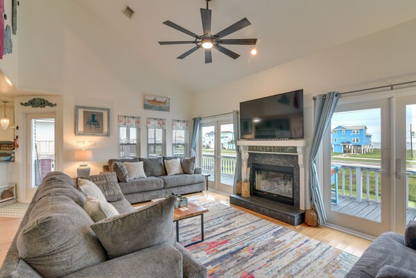 A spacious living room with a gray sofa set, a fireplace, a ceiling fan, and a large TV. - Pet-Friendly Condo w/ Seasonal Pool in Galveston! (Galveston)