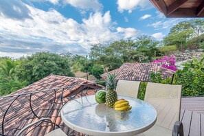Una mesa de vidrio redonda con una piña, sandía y plátanos, ubicada en una terraza con techo de tejas y rodeada de follaje y flores rosadas.