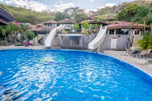 A swimming pool with two slides, surrounded by houses with red-tiled roofs, and a stone wall with a waterfall feature. - Hermosa Heights Villa 16 (Sardinal)