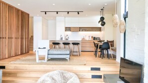 A modern living room with a sofa, ottoman, and coffee table. There is a dining area with a table and chairs. A kitchen with a bar area is visible in the background.