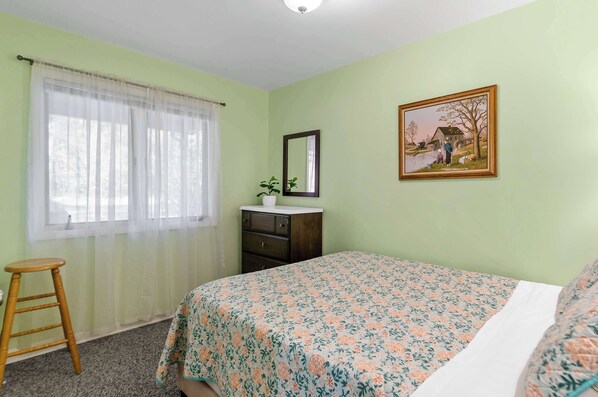 A bedroom with a floral bedspread, a wooden dresser, a mirror, and a painting on the wall.