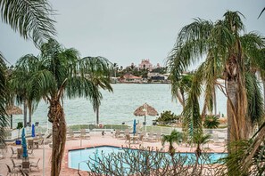 Zona de piscina con sillas de descanso y sombrillas, con vista a una masa de agua y edificios al fondo.