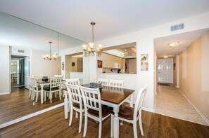 A dining area with a wooden table and white chairs, a chandelier, and a kitchen in the background.