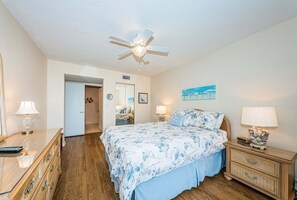 Un dormitorio con cama, mesitas de noche, ventilador de techo y una puerta que da a otra habitación.