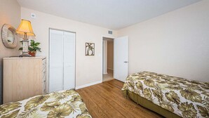 Un dormitorio con dos camas, un aparador de madera, una lámpara, una planta, una puerta y un cuadro colgado en la pared.