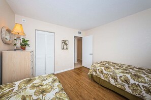 Une chambre avec deux lits, une commode en bois, une lampe, une plante, une porte et un tableau accroché au mur.