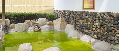 Um jardim japonês tradicional com lago para carpas, rodeado por pedras grandes e um muro de pedra.