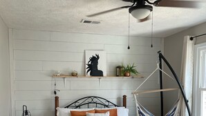 A bedroom with a bed, a hammock, a ceiling fan, a nightstand, and a wall-mounted shelf with decorative items. - Masters Rental Conveniently Located to Augusta National (Aiken)