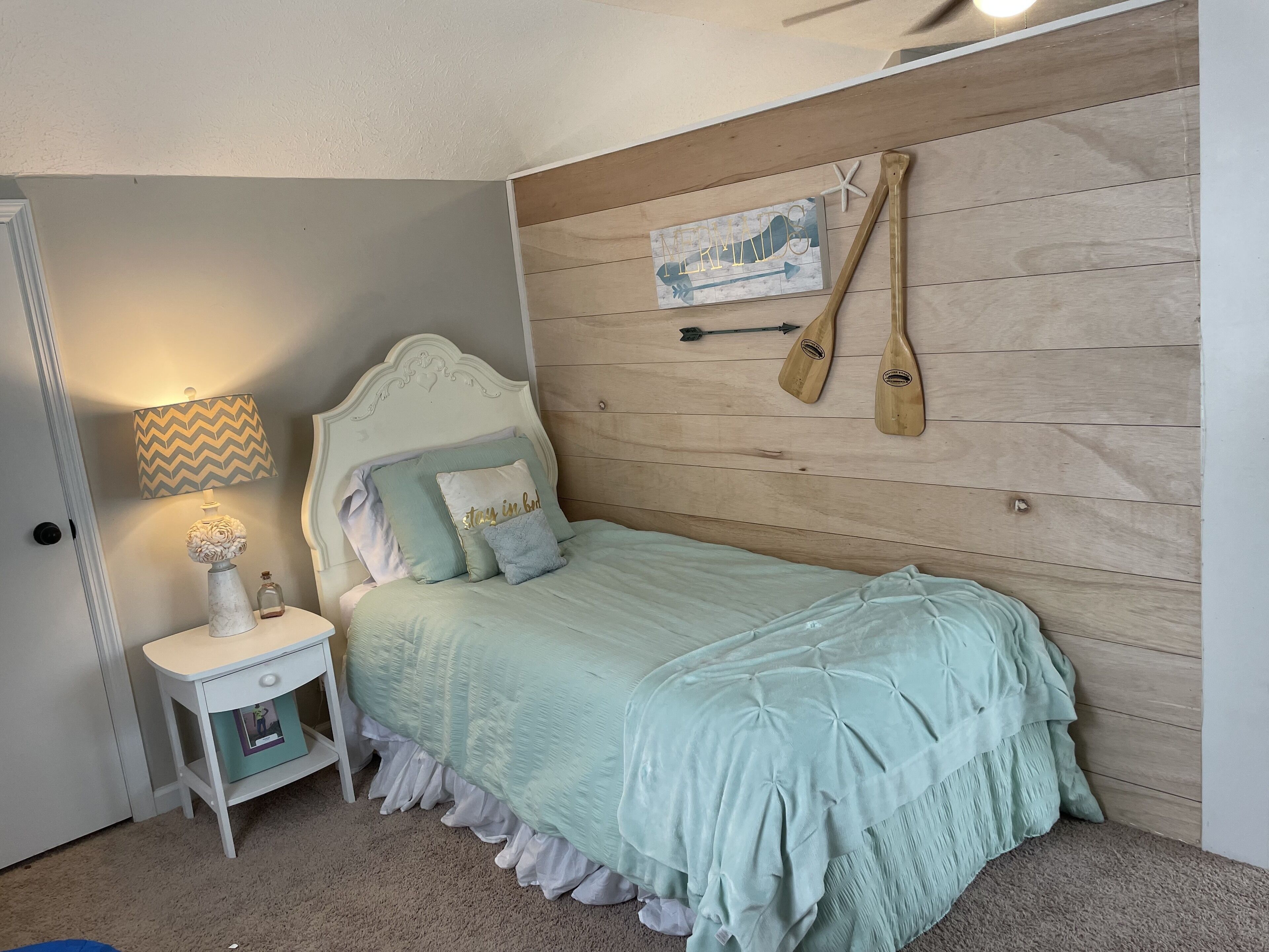 A bedroom with a bed, a nightstand, a lamp, a small table with a vase, and a wall with a wooden panel and two paddles.