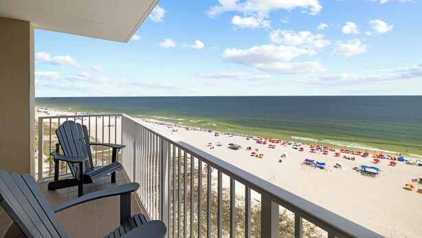 Un balcon avec deux fauteuils Adirondack donnant sur une plage parsemée de parasols et fréquentée par des baigneurs.