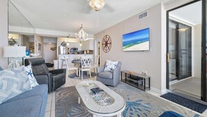Un salon moderne avec une table basse en verre, un canapé et un fauteuil gris. On y trouve également un coin repas avec une table et des chaises blanches, et une cuisine à l’arrière-plan.