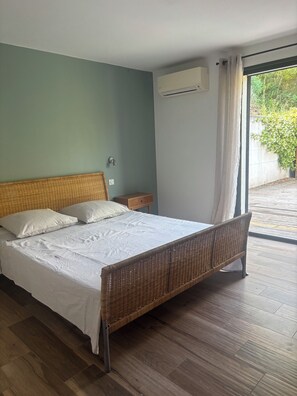 A bedroom with a bed, a nightstand, an air conditioning unit, and a view of the outdoors.