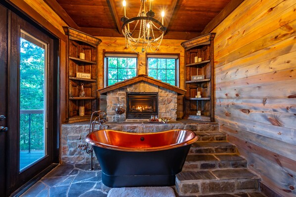 Une salle de bain rustique, aménagée dans une cabane en rondins, avec une baignoire sur pieds, une cheminée en pierre et des étagères en bois.