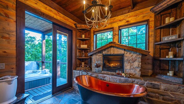 A rustic cabin interior with a stone fireplace, a large bathtub, and a cozy seating area with a hot tub. - Stunning copper tub room, outdoor Jacuzzi with view of Pine Lake (Broken Bow)