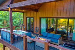 Une terrasse en bois avec un jacuzzi, des sièges d’extérieur et une porte vitrée donnant accès à un espace aménagé autour d’un foyer.