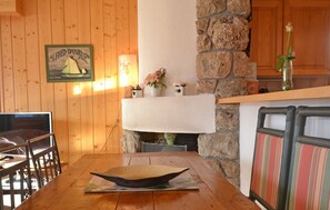 Ein Holzinnenraum mit Kamin aus Stein, einem Tisch mit Unterlage, Stühlen und einem gerahmten Bild an der Wand.