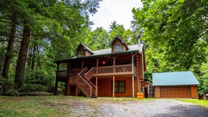 A log cabin with a balcony, a garage, and a gravel driveway surrounded by trees.