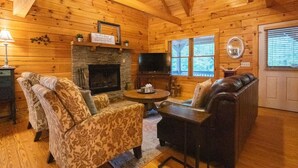 A cozy cabin interior with a fireplace, wooden walls, a television, and a seating area with patterned and leather sofas.