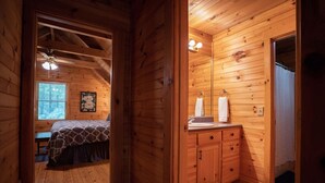 A wooden cabin bedroom with a bed, a window with blinds, a ceiling fan, and a bathroom with a sink and mirror.