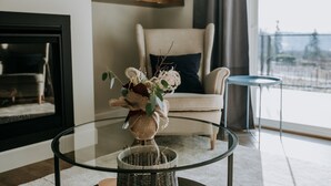 Un salon avec une table basse en verre, un panier en osier et un vase rempli de fleurs. On y trouve un fauteuil beige avec un coussin foncé, une petite table ronde et une cheminée.