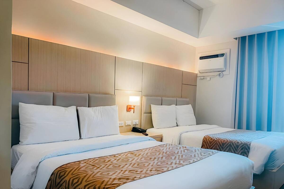 A hotel room with two beds, a ceiling fan, an air conditioning unit, and a window with curtains.