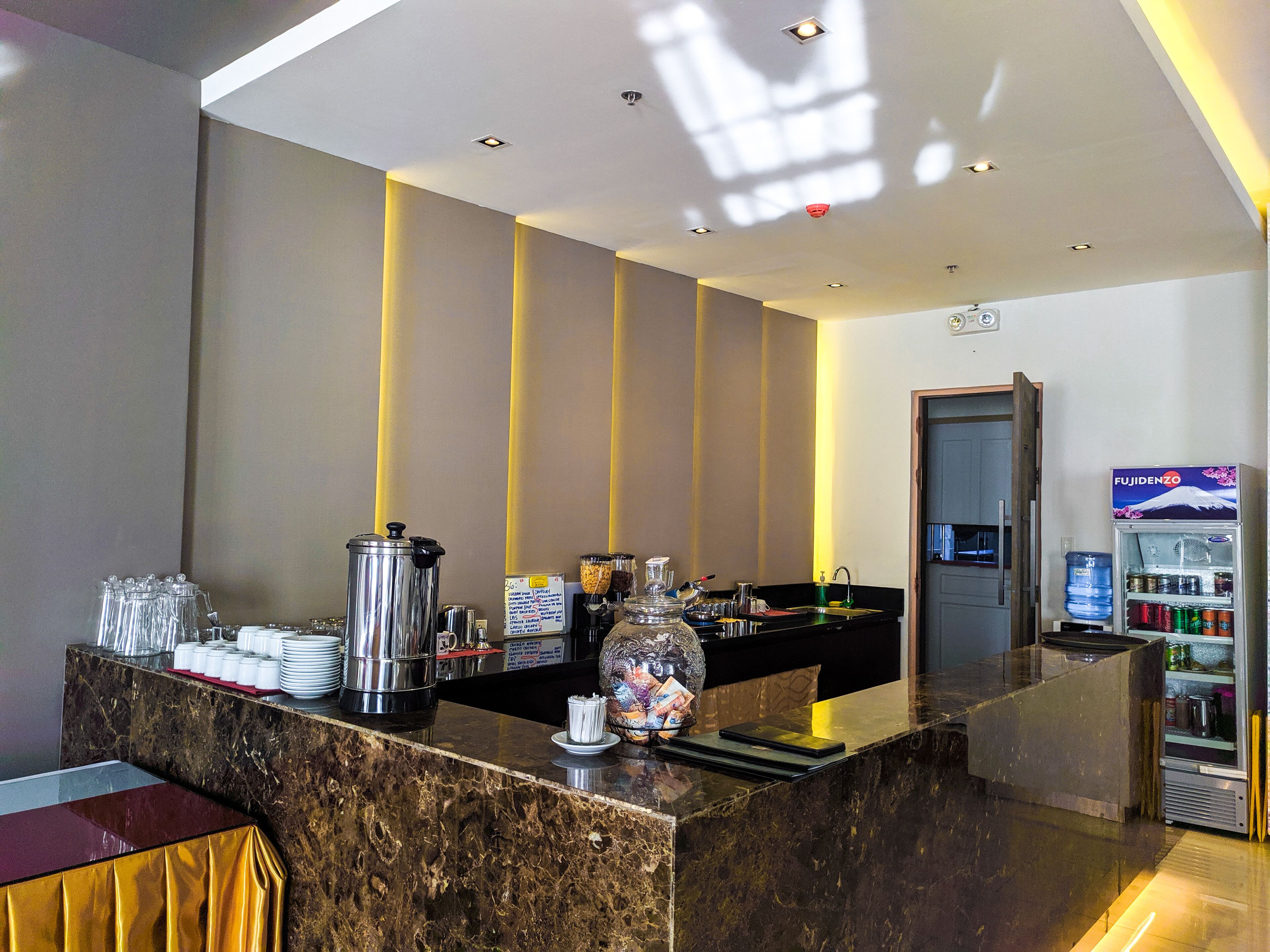 A hotel reception area with a marble counter, a coffee machine, a water dispenser, and a refrigerator stocked with beverages.
