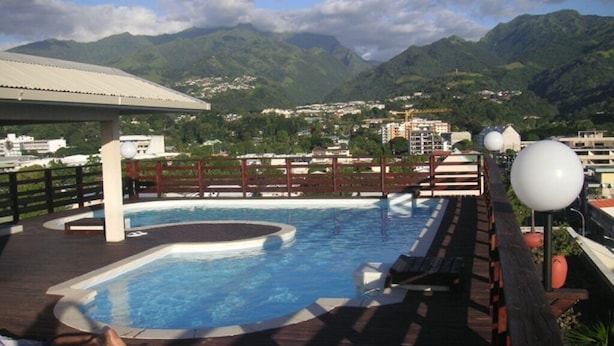 Ein Schwimmbecken mit Holzterrasse, umgeben von Bergen und Gebäuden.