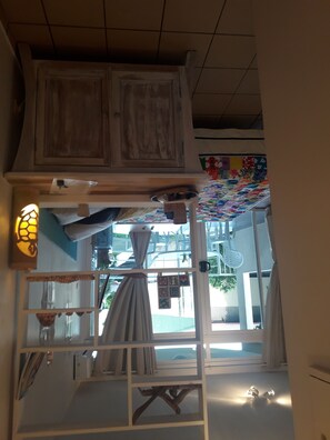 A room with a wooden cabinet, a white shelf, a colorful quilt, and a window with curtains. - A sweet and confortable studio downtown Papeete (Papeete)