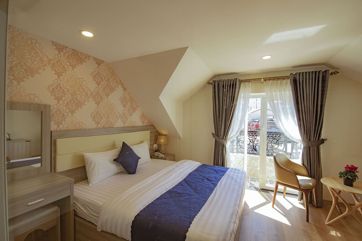 Quarto com cama, mesa de cabeceira, cadeira, pequena mesa com planta e varanda com cortinas.
