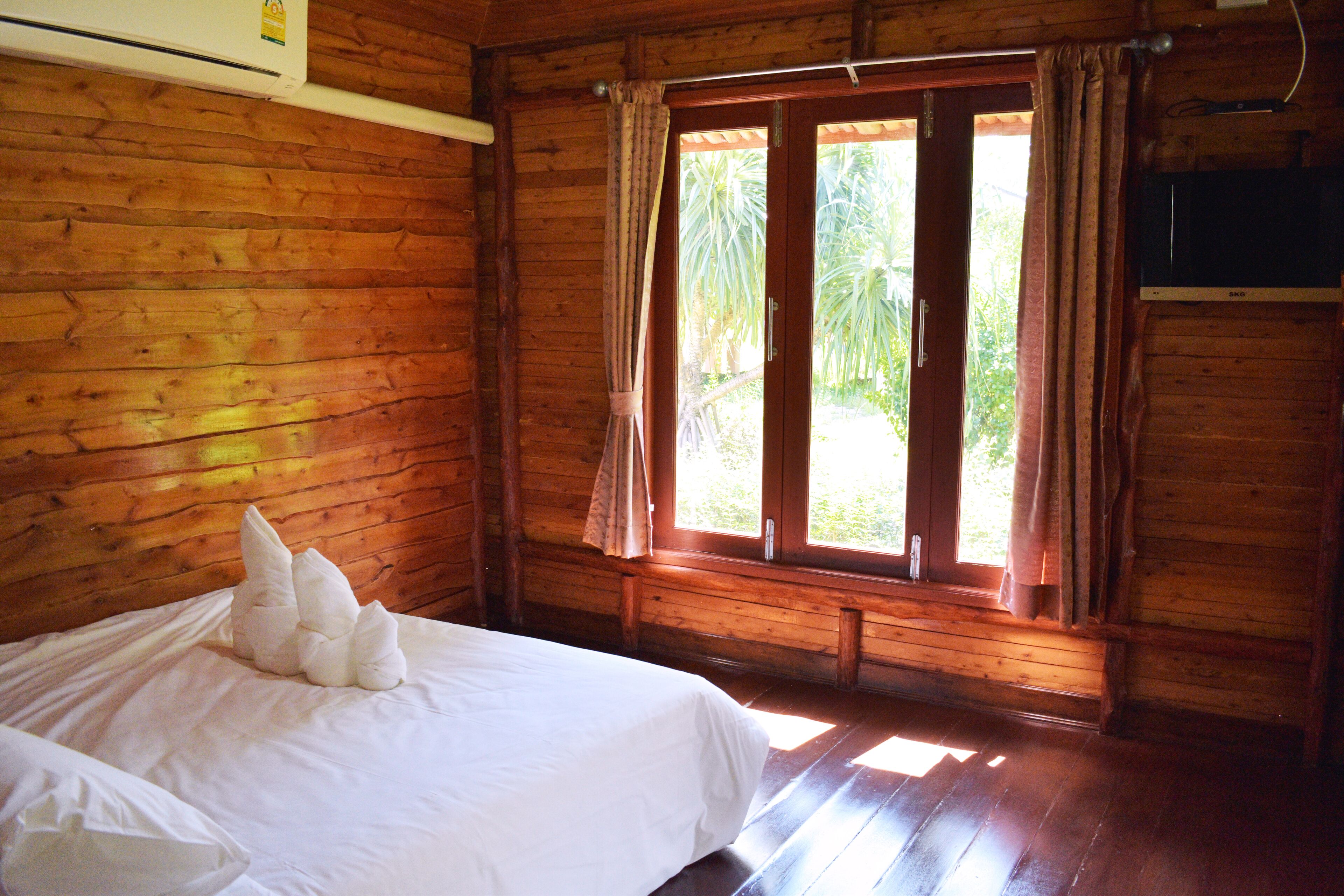 A wooden cabin room with a bed, a window with curtains, and a television on the wall.