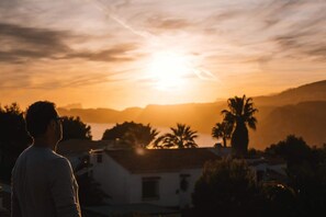 Une personne contemple un coucher de soleil avec vue sur un paysage côtier, comprenant des palmiers et un bâtiment doté d’un toit en pente.