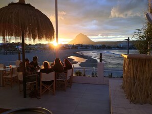 Une terrasse avec des sièges extérieurs, donnant sur une plage et une montagne, dotée d’un toit de chaume et offrant une vue sur le coucher de soleil.