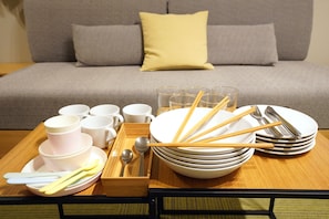 Una disposición para comer con recipientes, tazas, palillos y cubiertos sobre una bandeja, con un sofá y cojines al fondo.