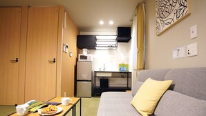 A compact hotel room with a kitchenette, a sofa, a small table with breakfast items, and a bed with a yellow pillow. - Koti Sopo Universal Bay 1 by Liaison (Osaka)