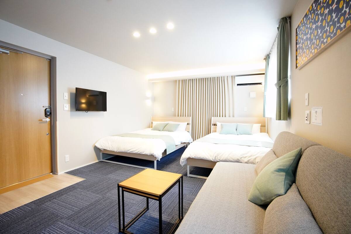 Habitación de hotel con dos camas, un sofá, una mesita, un televisor y una ventana con cortinas.
