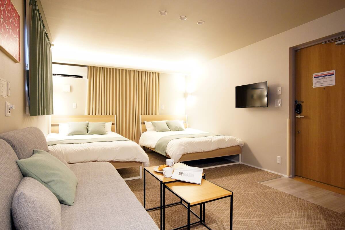 Habitación de hotel con dos camas, un sofá, una mesita con una taza y un libro, y un televisor en la pared.