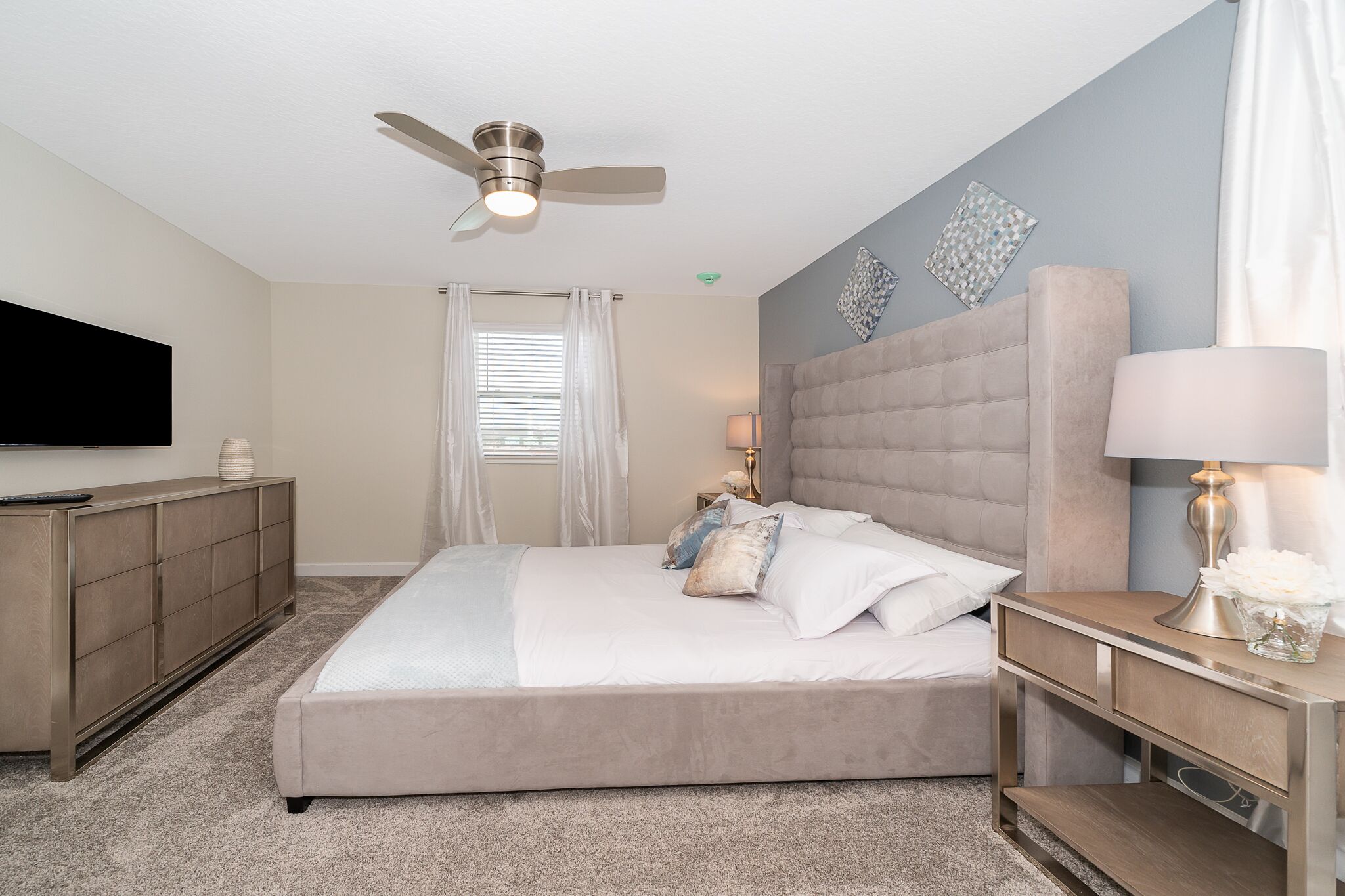 A bedroom with a large bed, a ceiling fan, a TV, a bedside table with a lamp, and a nightstand.