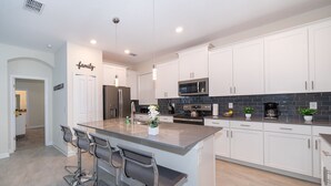 A modern kitchen with white cabinets, a central island with bar stools, stainless steel appliances, and a backsplash with dark tiles. - (WE2738) Storey Lake Home 5/5, Private Pool & Free Waterpark (Kissimmee)