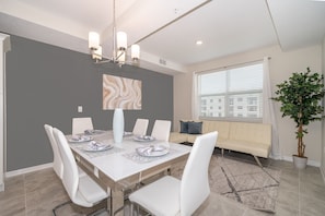 Un coin repas avec une table blanche dressée pour six personnes, un mur d’accent de couleur grise et une grande fenêtre avec des stores.